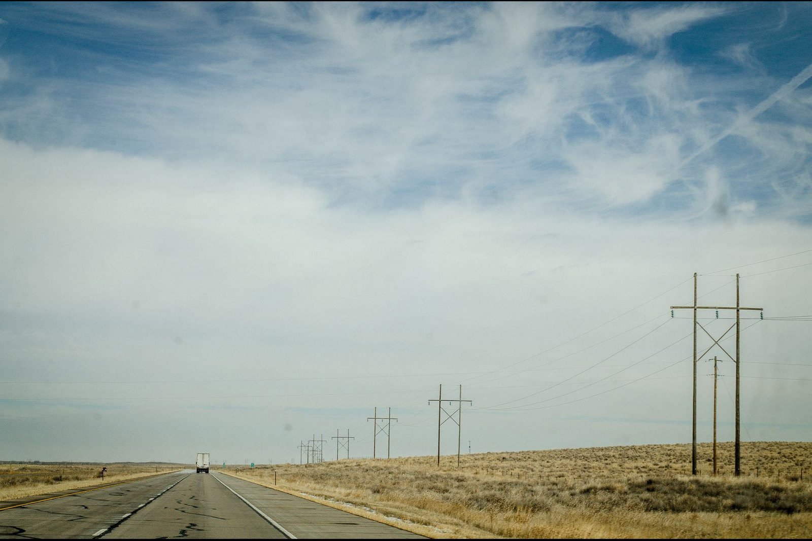 A rural highway and a line of wooden power poles crossing dry golden high desert rangeland under a wide sky in Arizona