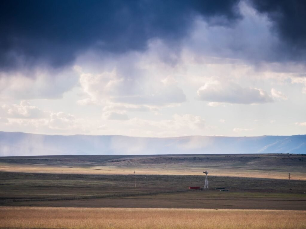 A lone windmill on open grassland in New Mexico under dark storm clouds, with a mesa on the horizon