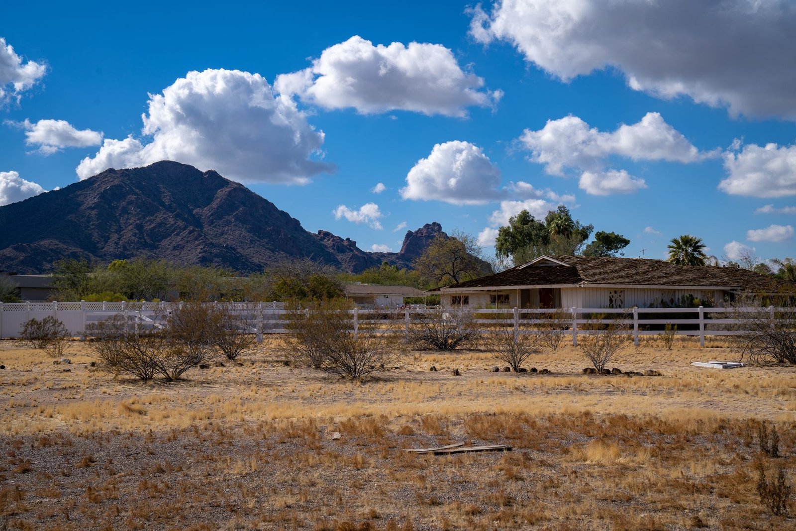 Rural Arizona land: a low ranch house behind a white rail fence, dry grassland in the foreground and a desert mountain range beyond