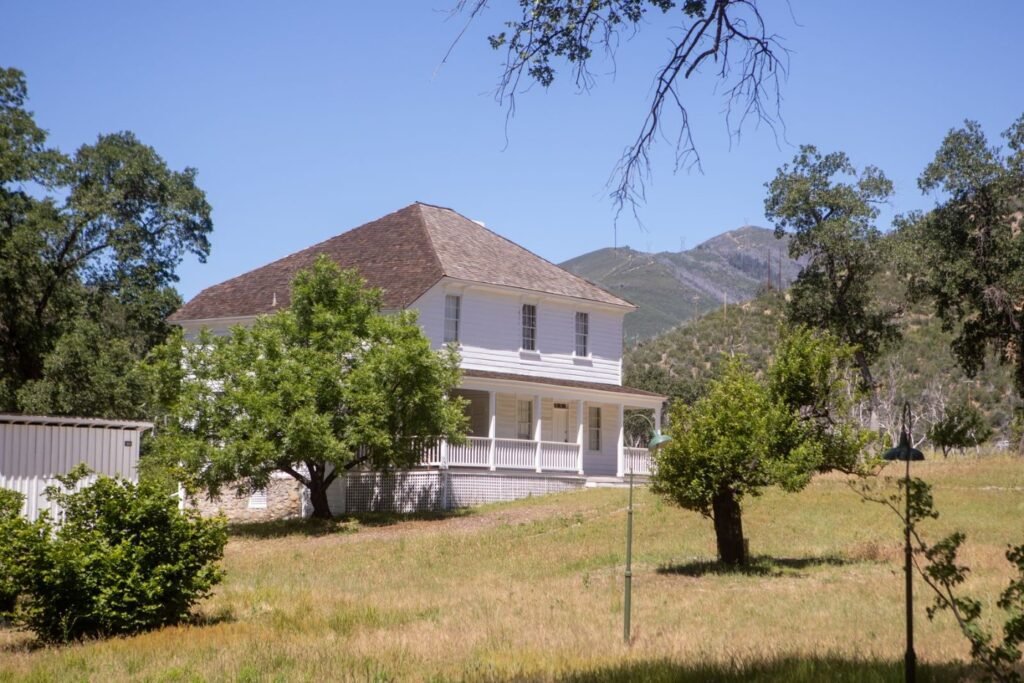 White two-story farmhouse surrounded by dry grass and oak trees in rural California