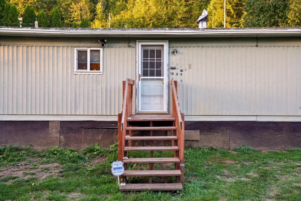 Single-wide manufactured home with metal siding, a white front door and wooden steps, set among trees